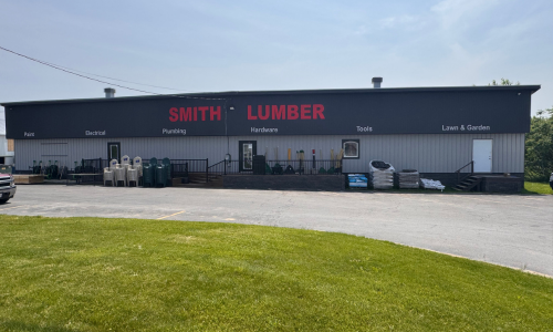 Smith Lumber storefront. The exterior is gray, with products like tanks and bags lined up outside. A paved parking area and a grassy lawn are in the foreground under a partly cloudy sky.