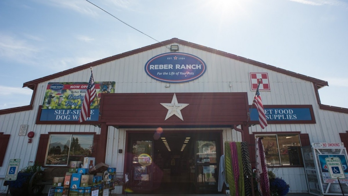 Front view of Reber Ranch, a pet supply store, under bright sunlight. The building is white with red trim, featuring American flags and a large star above the entrance.